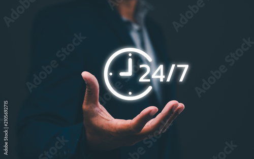 A person in a suit holds a glowing 24/7 clock icon, symbolizing continuous or round-the-clock service or availability.