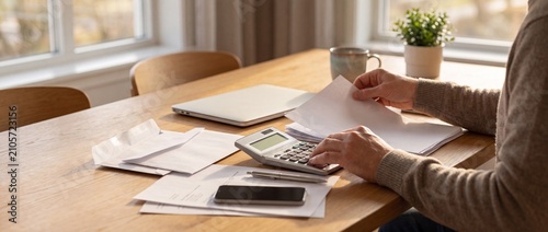 Person working with documents and calculator at a well lit wooden table in a cozy home office with cozy room large windows natural sunlight person organizing papers