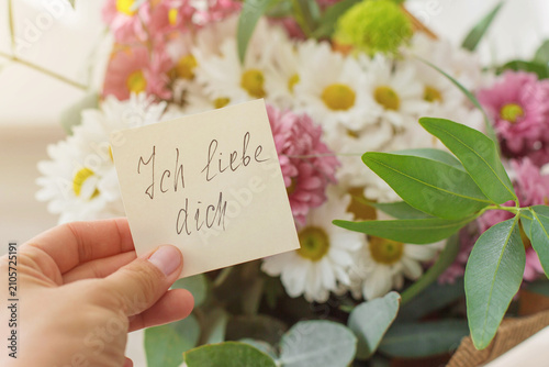 Hand holding note with handwritten German words Ich liebe dich I love you in english near bouquet of white daisies, pink chrysanthemums, eucalyptus leaves expressing romance, affection, heartfelt 