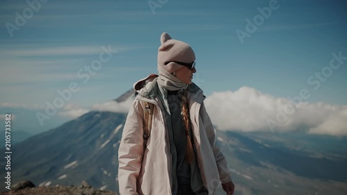 Young beautiful girl wearing sunglasses and carrying backpack stands against backdrop of majestic volcano and admiring surroundings from above. Slow motion