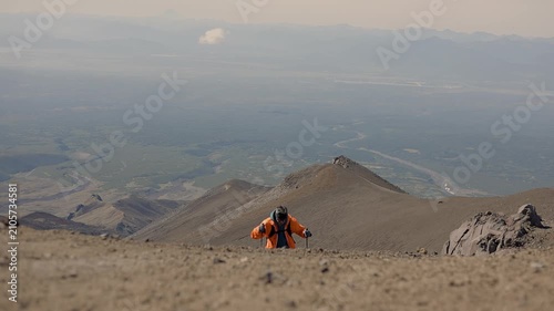 Kamchatka, Russia - August 24, 2025: Young man with mustache and orange jacket climbs ridge using trekking poles against backdrop of majestic volcano with snow-capped peak.