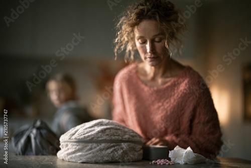 Woman in pink sweater packs lunch in kitchen, bread loaf, metal lunch container, and crumpled paper on counter as child watches