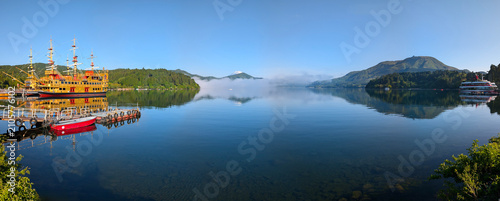 the calm, smooth waters of Lake Ashi with morning fog on the surface and boats surrounded by forested mountains and overlooking Mount Fuji with its snow-capped peak against a blue, cloudless sky