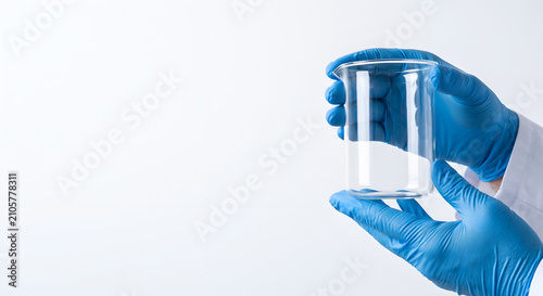 Scientist hands in blue gloves holding empty clean laboratory beaker against white.