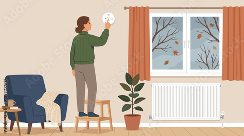 Woman standing on stepladder adjusts a smart thermostat in a cozy room during autumn. Concept of energy efficiency and climate control at home.