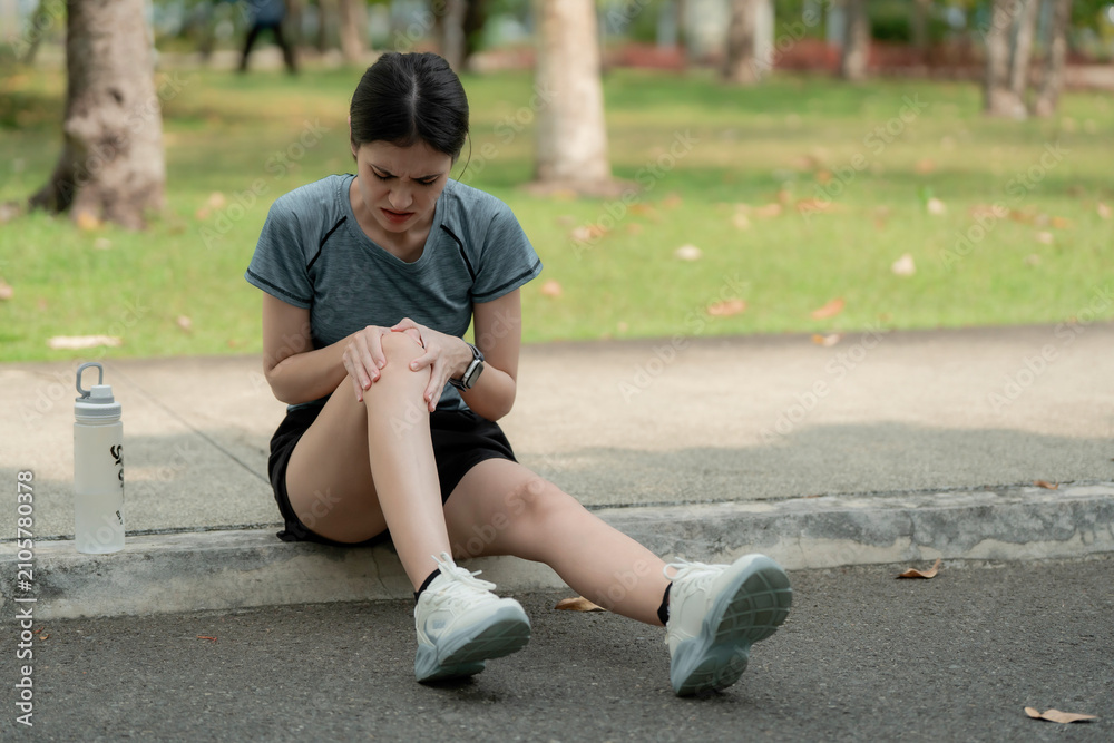 Fototapeta premium Young woman sitting on curb with knee pain after running in park, holding injured joint with discomfort, sports injury and overuse strain fitness concept. knee, pain, joint, strain, muscle,