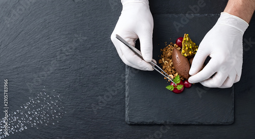 Chef using fine tweezers to decorate gourmet dessert on black stone plate dish.