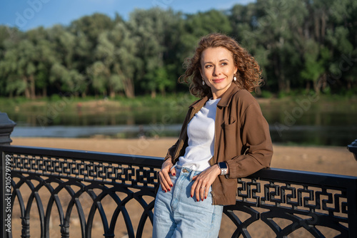 Curly-haired woman leaning on a fence by the river, sunny day.