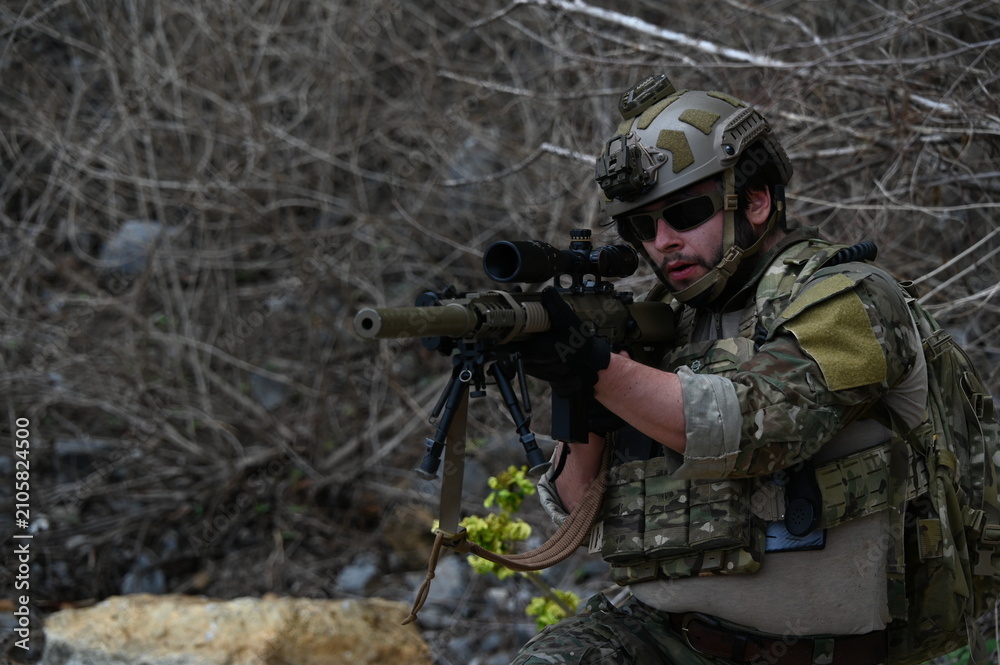 Fototapeta premium Tactical operator in camouflage gear rests with a rifle on rocky terrain during outdoor field training. Concept of military exercise, security, readiness, and tactical operations.
