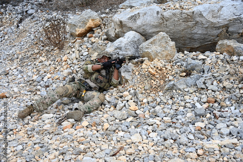 Obraz premium Focused military infantry soldier lying prone on rugged rocky terrain aiming a tactical assault rifle during a dangerous combat training mission for national defense and special security.