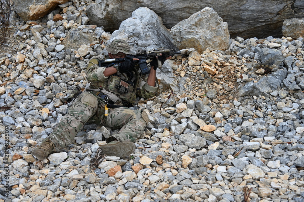 Obraz premium Focused military infantry soldier lying prone on rugged rocky terrain aiming a tactical assault rifle during a dangerous combat training mission for national defense and special security.