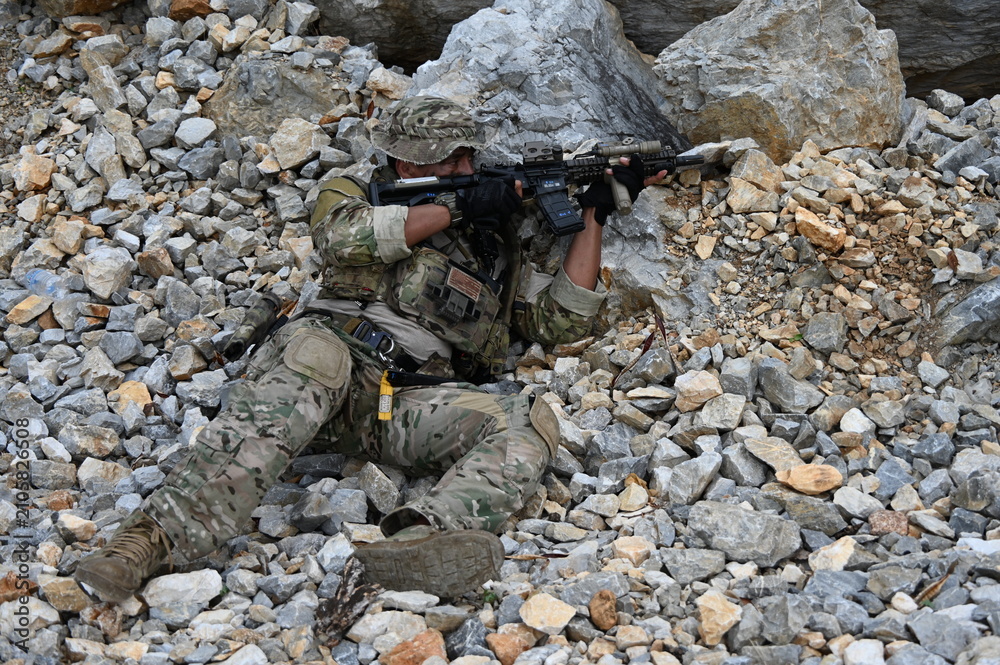 Obraz premium Focused military infantry soldier lying prone on rugged rocky terrain aiming a tactical assault rifle during a dangerous combat training mission for national defense and special security.