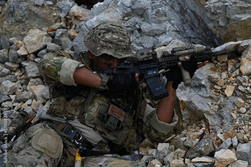 Obraz premium Focused military infantry soldier lying prone on rugged rocky terrain aiming a tactical assault rifle during a dangerous combat training mission for national defense and special security.