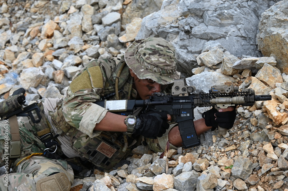 Obraz premium Focused military infantry soldier lying prone on rugged rocky terrain aiming a tactical assault rifle during a dangerous combat training mission for national defense and special security.