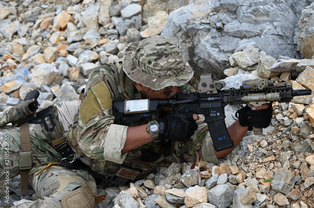 Obraz premium Focused military infantry soldier lying prone on rugged rocky terrain aiming a tactical assault rifle during a dangerous combat training mission for national defense and special security.