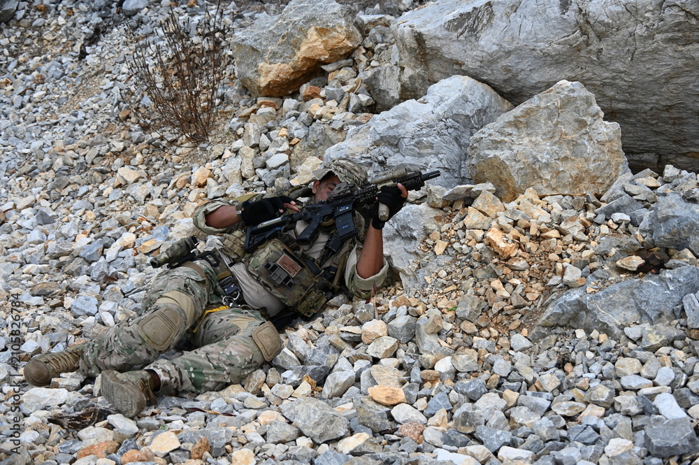 Obraz premium Focused military infantry soldier lying prone on rugged rocky terrain aiming a tactical assault rifle during a dangerous combat training mission for national defense and special security.