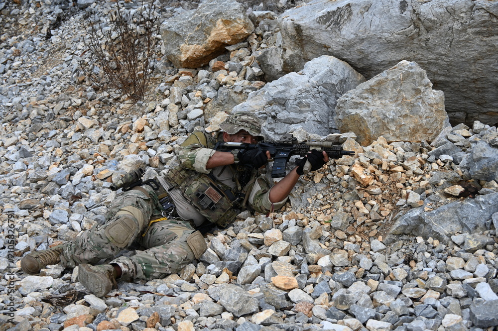 Obraz premium Focused military infantry soldier lying prone on rugged rocky terrain aiming a tactical assault rifle during a dangerous combat training mission for national defense and special security.