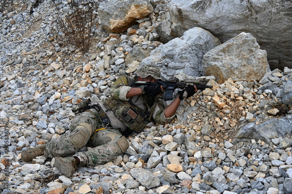 Obraz premium Focused military infantry soldier lying prone on rugged rocky terrain aiming a tactical assault rifle during a dangerous combat training mission for national defense and special security.