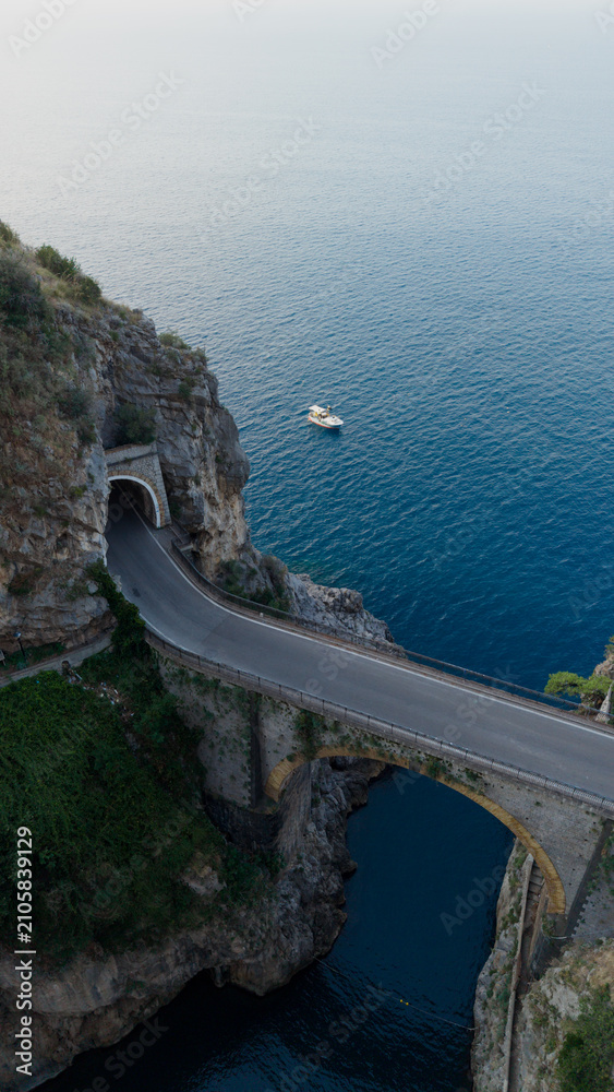 Obraz premium Fiordo di furore bridge and coastal road tunnel connecting rugged cliffs, overlooking the tranquil mediterranean sea with a boat gently floating in the clear blue water, amalfi coast, italy