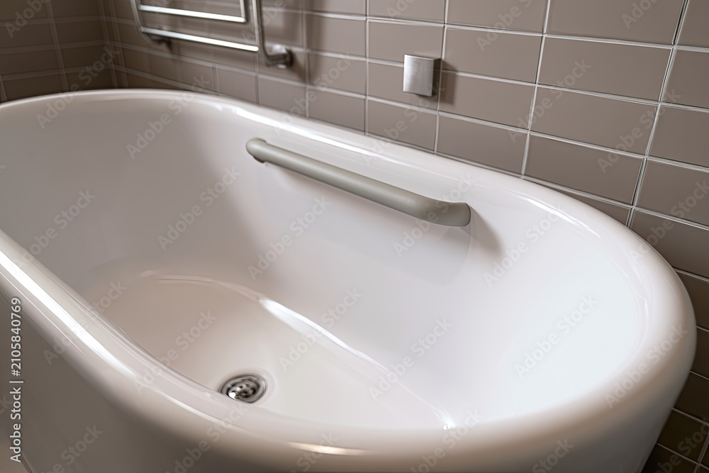 Fototapeta premium A clean white bathtub with a chrome drain and silver grab bar against tiled walls