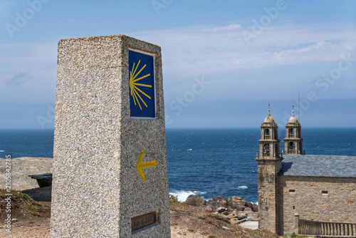 Camino de santiago waymarking stone with shell symbol and yellow arrow indicating the route, overlooking a coastal church and the atlantic ocean