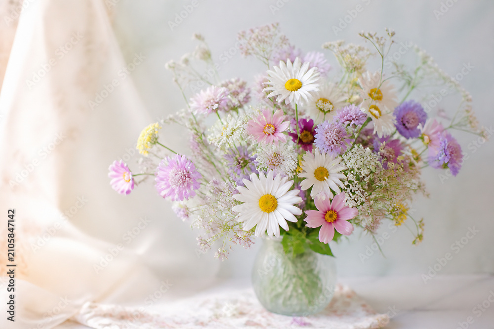 Obraz premium A bouquet of mixed wildflowers in a glass vase sits on a light surface near draped fabric.