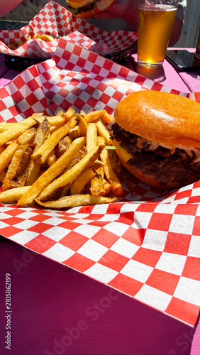 A hamburger topped with brisket and served with fries, in a basket with a red a white checked liner. Seved outside on a picnic table.