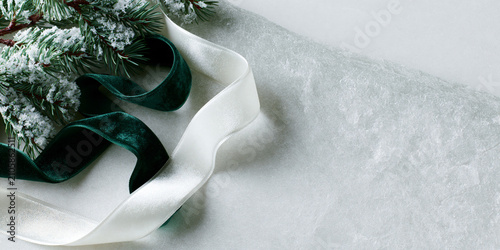 Elegant snowy pine branch with emerald green velvet and white satin ribbons on frosted textured background