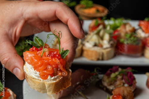 Obraz na plátne A hand picks up a small salmon bruschetta from a platter filled with various gourmet tapas appetizers against a dark background