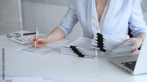 Businesswoman managing heavy workload, organizing financial documents for corporate accounting, tax preparation, and audit, working at office desk with stack of paper files and laptop. Audit and taxes