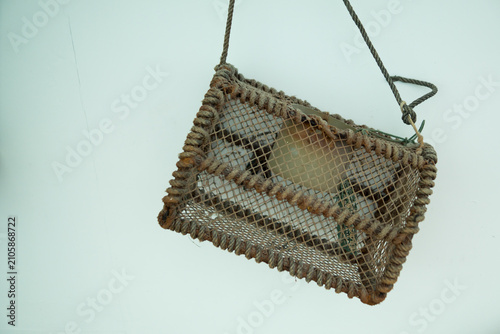 A rustic handmade fishing cage reinforced with thick brown rope, used for commercial or artisanal trapping of seafood, hanging on a white wall.