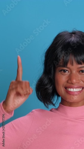 A cheerful young Southeast Asian woman with short black hair and bangs smiles widely while pointing her index finger upwards, presenting a great idea over a solid blue studio background.