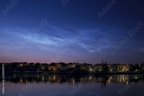 The outline of buildings of city over lake in night scenery. Silvery clouds NLC in the sky.	