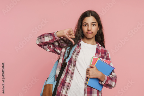 Foto Displeased schoolgirl with backpack and books in hands showing thumbs down on pi