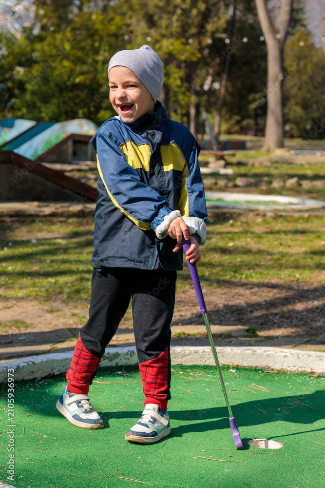 Obraz premium A young boy swings a mini golf club toward a yellow ball on an outdoor putting green, with trees, a bench and a gazebo visible in the sunny park background