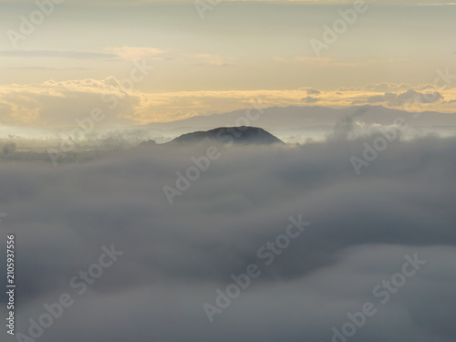 A misty morning view of Mt Wellington emerging from thick fog in Auckland, New Zealand. The sun attempts to break through the clouds, creating a serene and atmospheric landscape.