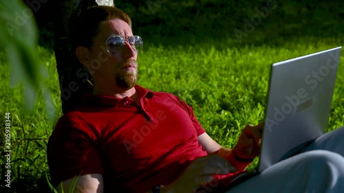 Busy man working on a laptop while sitting on the grass in a city park sitting under tree.