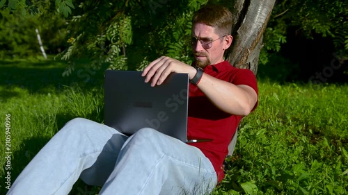 Busy man working on a laptop while sitting on the grass in a city park sitting under tree.