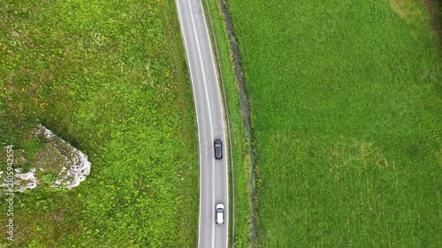Aerial view of a black car driving along a winding rural road surrounded by lush green trees and rocky cliffs. Travel by auto at scenic landscape. Vehicle journey through natural beauty of countryside