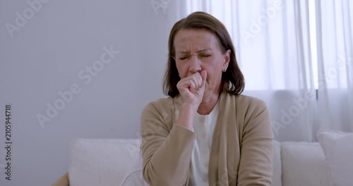 Senior woman sits on living room sofa, holding chest and repeatedly coughing into hand, expressing sudden throat discomfort, health concern, respiratory symptom, and need for caring support at home.