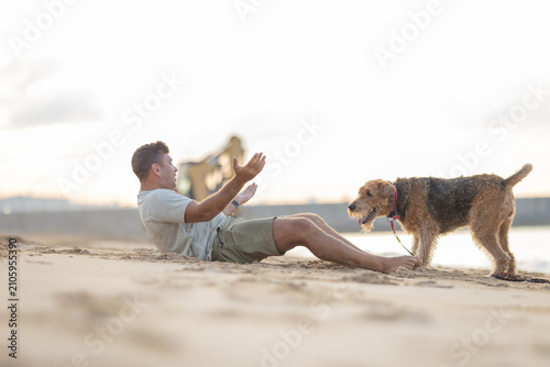 Fotografie Male dog owner sitting on sandy beach interacts with his pet Airedale Terrier during sunset, with water and distant figures in the background