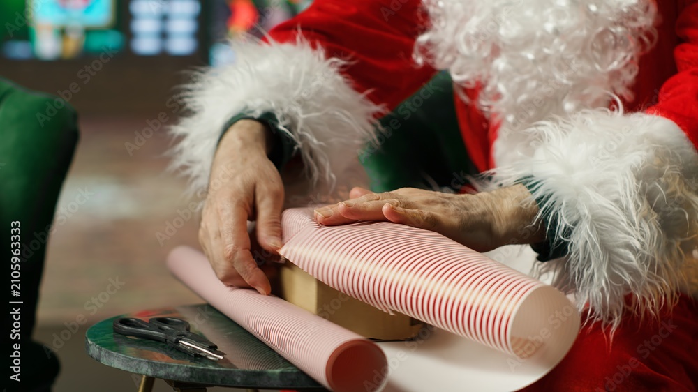 Fototapeta premium Santa Claus wraps presents in colorful paper after finishing the nice list and prepares the gifts for his Christmas journey to visit children.