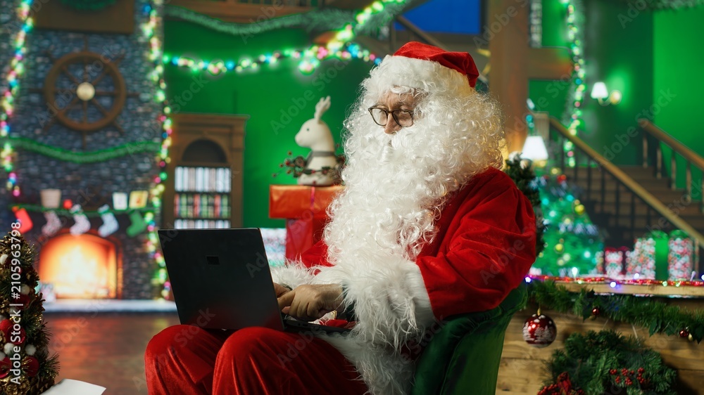 Fototapeta premium A smiling man dressed as Santa Claus reads a letter from children on a laptop and reviews their Christmas wish lists while wearing a cheerful red costume in a festive holiday setting.