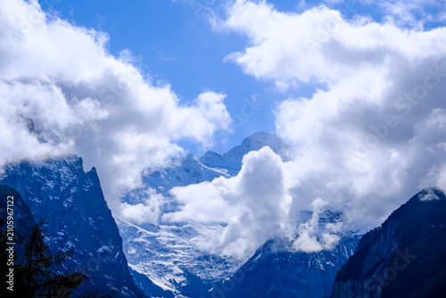 A stunning, high-altitude landscape photograph looking south from the alpine village of Wengen down into the iconic Lauterbrunnen Valley. The snow-shrouded peak of the massive Jungfrau mountain emerge