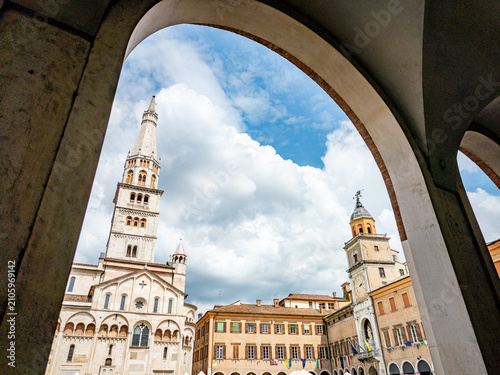 Modena Cathedral and Ghirlandina Tower rising above Piazza Grande, framed by elegant arched porticos, showcasing historic Romanesque architecture under a blue sky with clouds