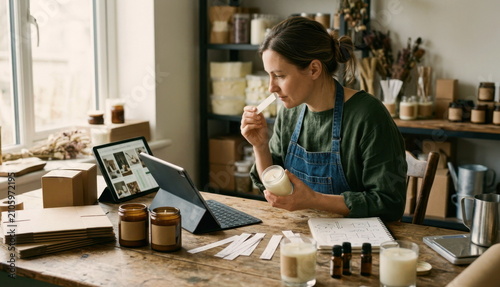 Artisan candle maker comparing seasonal product ideas with tablet, scent strips, kraft boxes, and sample jar candles in a small studio