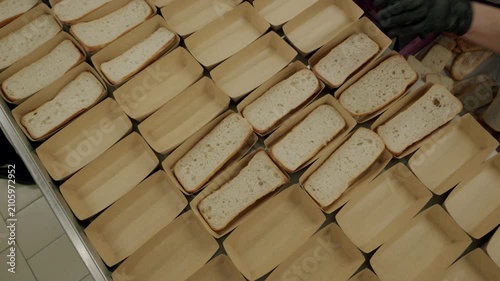 Worker in gloves places sliced bread into paper trays on a table, showcasing a systematic arrangement of loaves in a commercial kitchen environment
