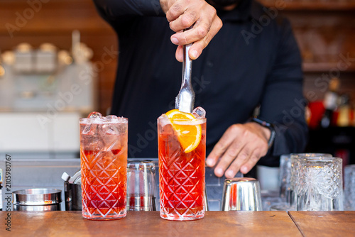 Bartender adding an orange slice to two red cocktails in highball glasses on a bar counter
