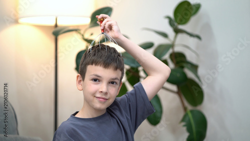 Young boy smiles while using a head massager on his scalp at home. Cozy indoor scene with soft light and plant.