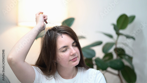 Young woman with closed eyes enjoys a soothing head massage using a metal wire massager, promoting deep relaxation and stress relief in a calm home setting with soft light.